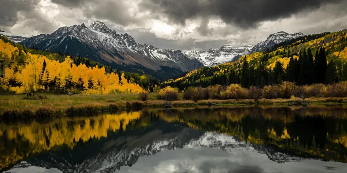 Snow-capped mountains and golden autumn trees reflected in a still lake under stormy skies