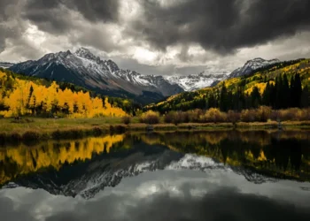 Snow-capped mountains and golden autumn trees reflected in a still lake under stormy skies