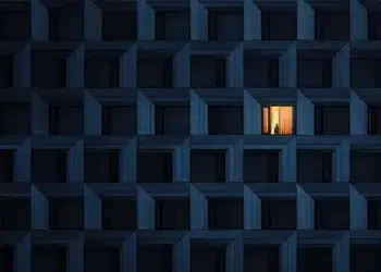 A solitary figure stands in the only lit window of a dark apartment building facade at night
