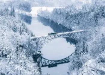 Train crossing a steel arch bridge over a frozen river in a snowy forest