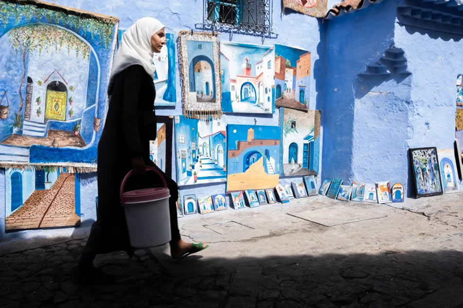 Muslim Lady walking with bucket backdrop of blue color wall with full of artistic frames Morocco - Captivating Street Photography by Giancarlo Zuccarone