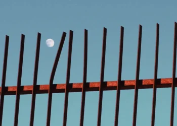 Full moon visible behind rusty iron fence against blue sky