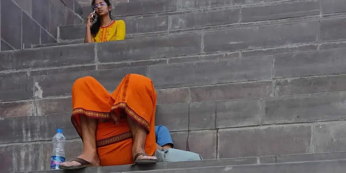 Man in orange dhoti and woman in yellow kurta on phone sitting on stone ghat steps, street photographers