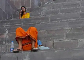 Man in orange dhoti and woman in yellow kurta on phone sitting on stone ghat steps, street photographers