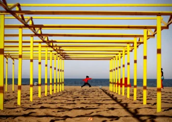 A child kicks a ball on a beach framed by yellow wooden structures