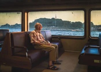 Elderly man sitting alone on a ferry bench, gazing out the window at water and distant shoreline
