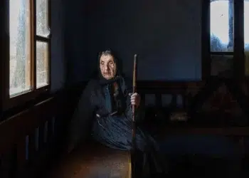 Elderly woman sitting by window holding a wooden stick in dim light