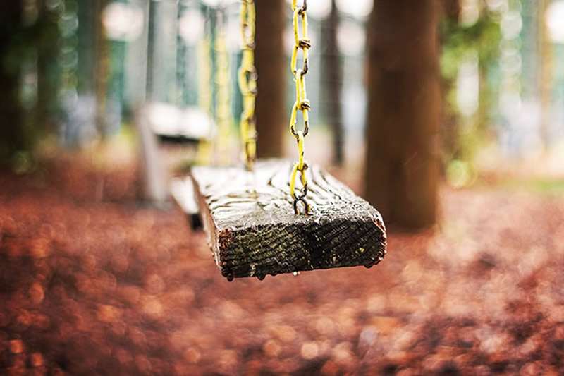 Macro-style photo of a wet wooden swing hanging by chains, with rain falling and autumn leaves softly blurred in background.