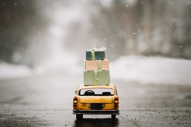 Macro photo of a miniature yellow car carrying gift boxes on a snowy road, with soft falling snow and blurred background.