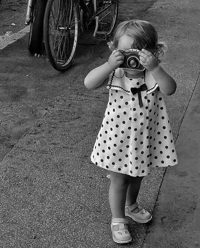 Timeless and Emotional: 30 Black and White Photos That Touch the Soul 32 Black and white photo of a small child wearing a polka dot dress holding a vintage camera up to her face while standing on a street near a bicycle, capturing a moment with innocent curiosity.