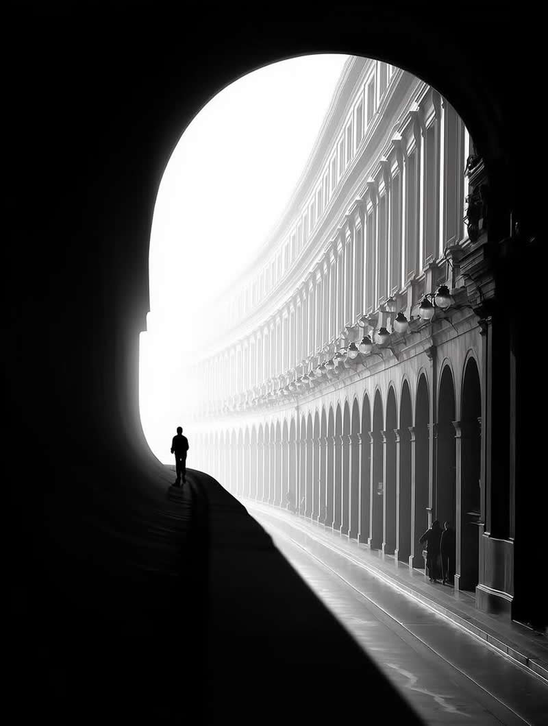 Timeless and Emotional: 30 Black and White Photos That Touch the Soul 33 Black and white photo of a lone person walking through a dark tunnel toward a bright arched opening, revealing a long row of classical buildings and arches outside, creating a dramatic contrast of light, shadow, and perspective.