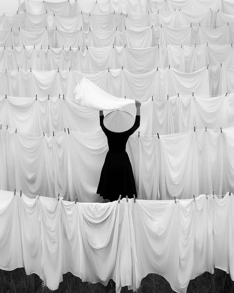 Timeless and Emotional: 30 Black and White Photos That Touch the Soul 34 Black and white photo of a person wearing a conical hat standing among rows of hanging white sheets on clotheslines, lifting a cloth overhead, creating a striking pattern of repeating fabric textures and minimalist contrast.