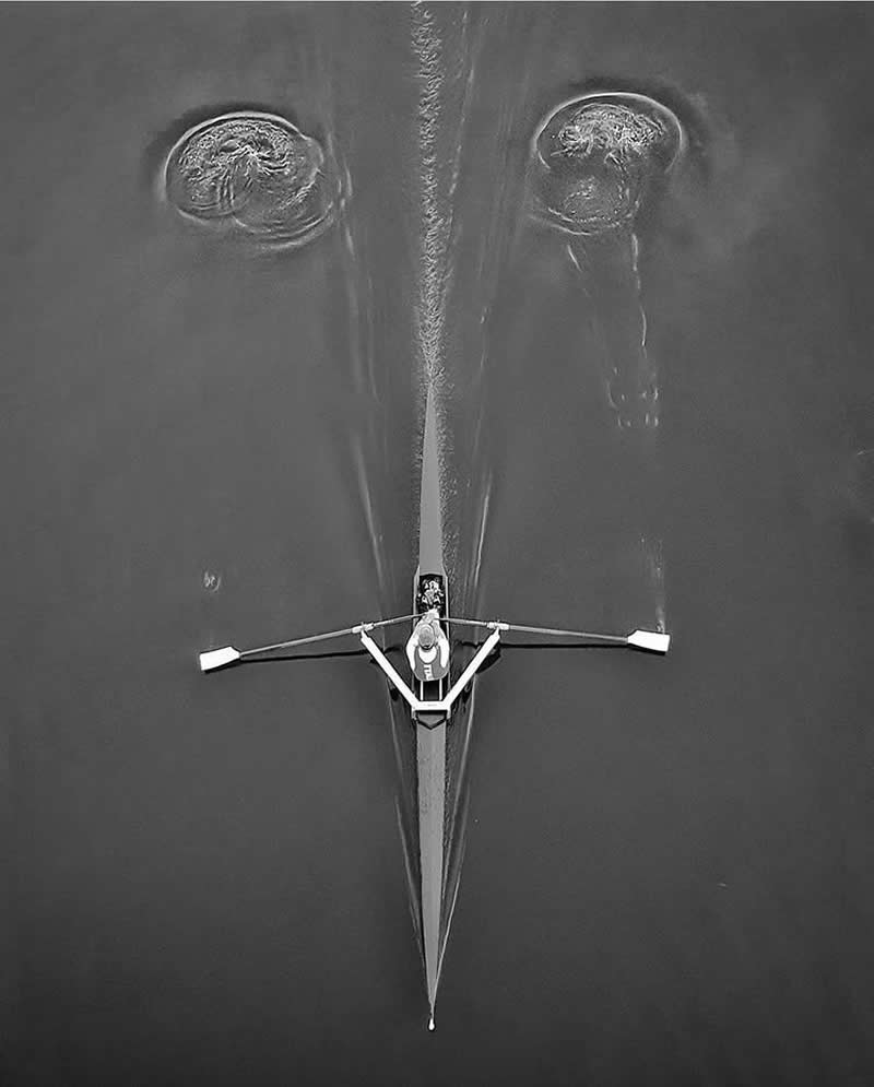 Timeless and Emotional: 30 Black and White Photos That Touch the Soul 38 Black and white aerial photo of a lone rower gliding across calm water, with oars creating symmetrical ripples that resemble eyes, forming a striking minimalist composition.