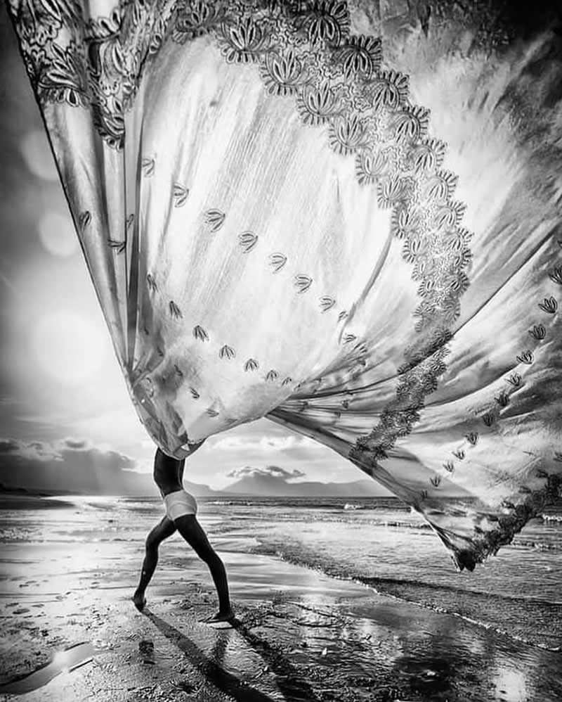 Timeless and Emotional: 30 Black and White Photos That Touch the Soul 39 Black and white photo of a person walking along a wet shoreline while holding a large flowing embroidered fabric above their head, the cloth billowing dramatically in the wind against a bright sky and reflective beach.
