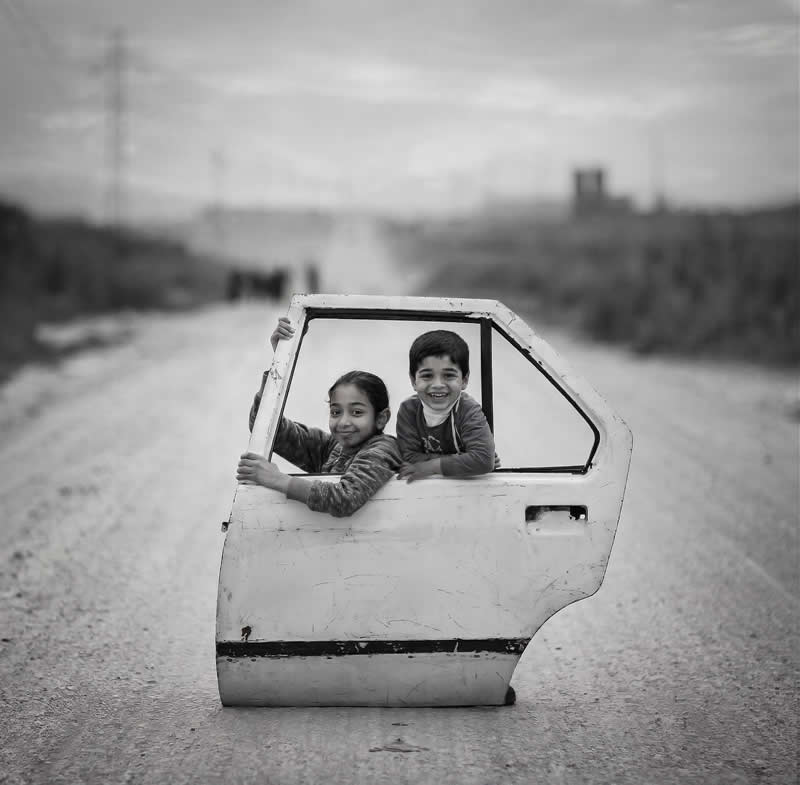 Timeless and Emotional: 30 Black and White Photos That Touch the Soul 46 Black and white photo of two smiling children pretending to ride in a car using a detached car door placed on a quiet rural road, capturing a playful moment of childhood creativity and joy.