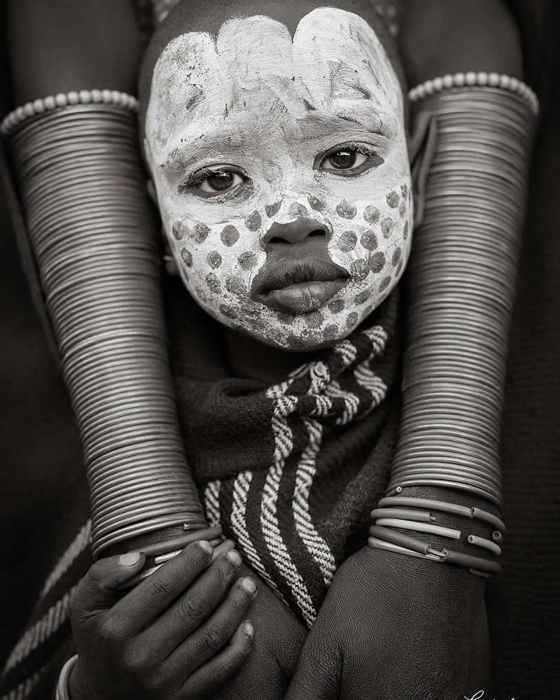 Timeless and Emotional: 30 Black and White Photos That Touch the Soul 51 Black and white portrait of a young child with traditional face paint featuring white patterns and dots, framed by an adult’s arms adorned with stacked metal bracelets, highlighting cultural identity and intimate human connection.
