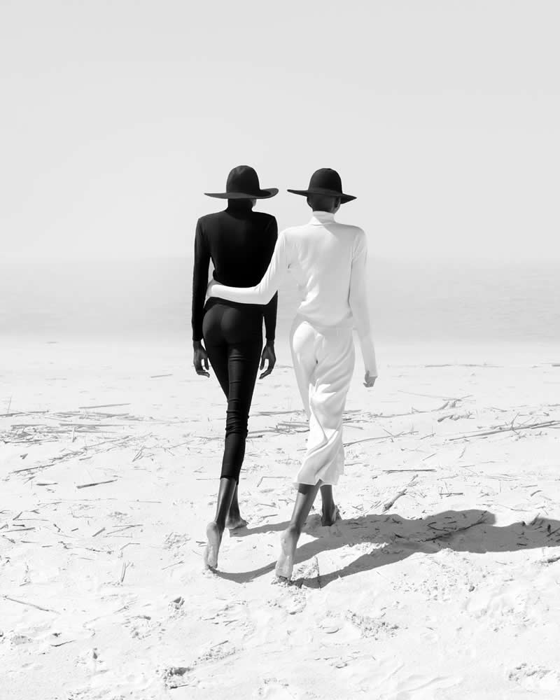 Timeless and Emotional: 30 Black and White Photos That Touch the Soul 56 Black and white minimalist photo of two people walking barefoot across a bright sandy landscape, one dressed in black and the other in white, both wearing wide-brim hats and walking side by side.