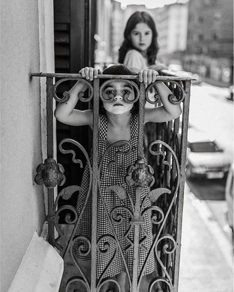 Timeless and Emotional: 30 Black and White Photos That Touch the Soul 61 Black and white photo of a young child standing behind an ornate balcony railing, holding the bars and looking out toward the street, while another child stands slightly blurred in the background.
