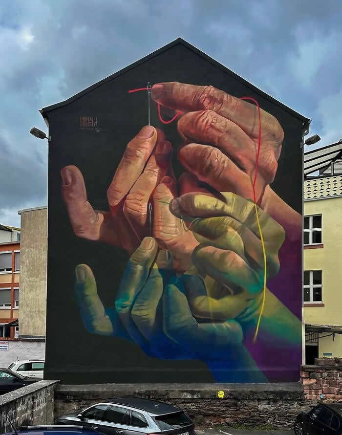 A large mural on a building wall depicts multiple realistic hands in different skin tones carefully threading a needle with colorful string, symbolizing unity, diversity, and human connection in an urban setting.