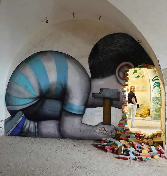 A creative street mural of a child appearing to break through a wall with a hammer, with colorful bricks scattered on the ground, blending illusion and reality as a person walks through the archway in an urban setting. Neuf-Brisach, France.