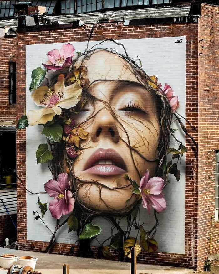 A large hyper-realistic mural of a woman’s face intertwined with vines and blooming flowers, painted on a brick wall, blending human beauty with nature in a striking and surreal urban street art composition.