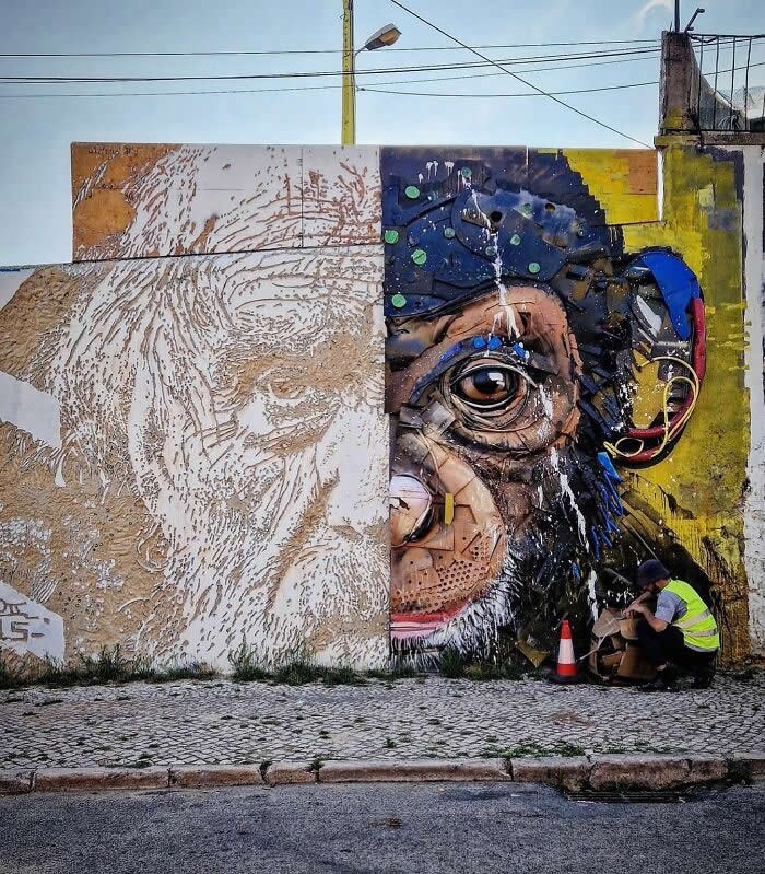 A striking mural split into two halves shows a rough sketch of an elderly man transitioning into a detailed, colorful chimpanzee face, with a worker sitting nearby, emphasizing the transformation from concept to finished street art. Lisbon, Portugal