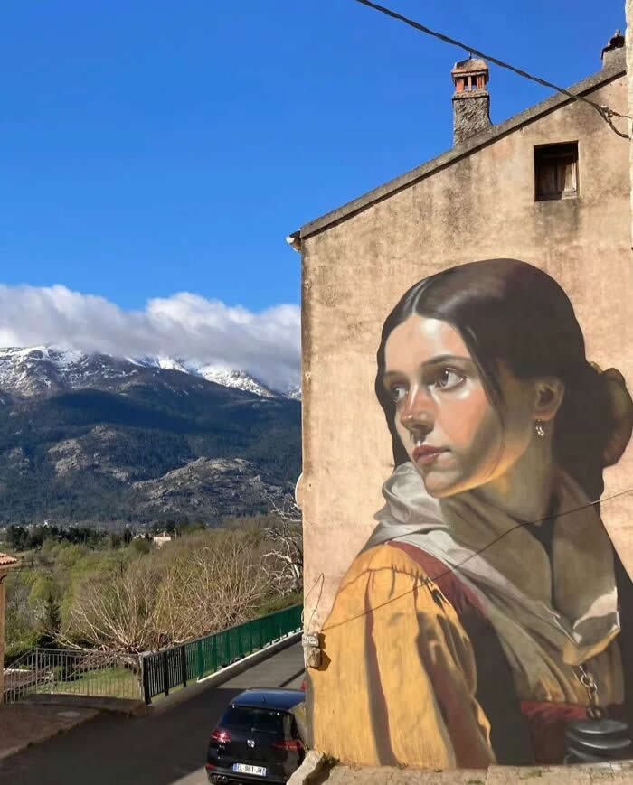A realistic mural of a young woman painted on the side of a rustic building, her face turned thoughtfully toward distant snow-covered mountains, blending art seamlessly with the surrounding natural landscape and quiet street below. Lozzi, Corsica.