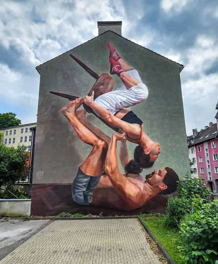 A large mural of two acrobats balancing in a dynamic pose, one lifting the other upside down with a chair, painted on a building wall, showcasing strength, trust, and motion against an urban neighborhood backdrop. Wuppertal, Germany.