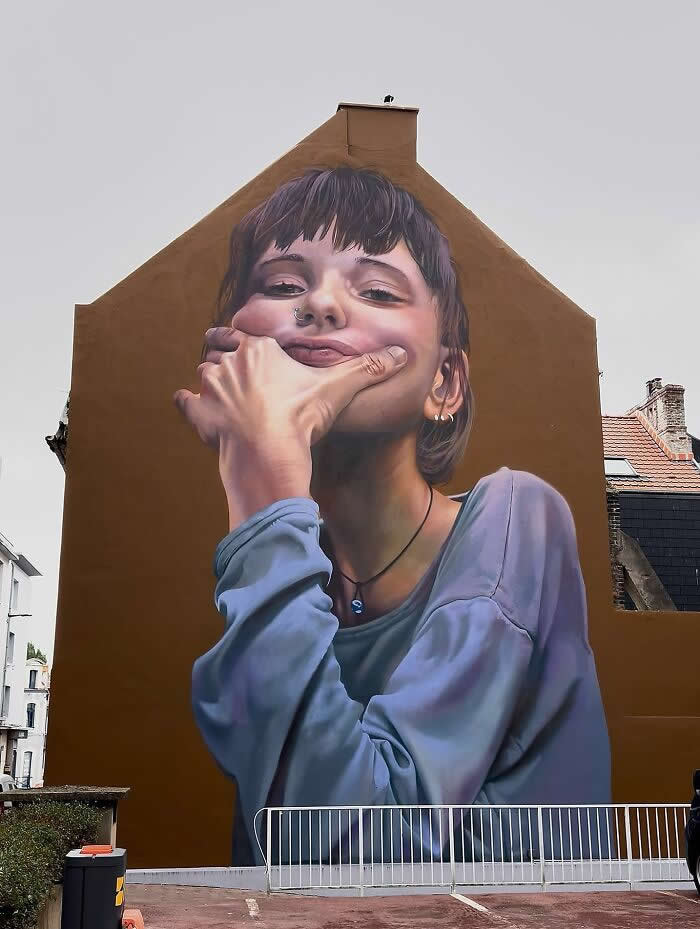 A hyper-realistic mural of a young person leaning forward with a playful, defiant expression, resting their chin on their hand, painted on a large building wall in an urban neighborhood. Boulogne-sur-Mer, France.