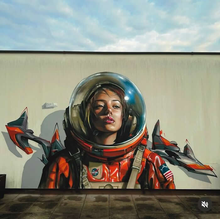 A large street mural of a female astronaut in a reflective helmet and orange space suit, with stylized spacecraft elements behind her, painted on an urban wall under a cloudy sky. Prague, Czech Republic.