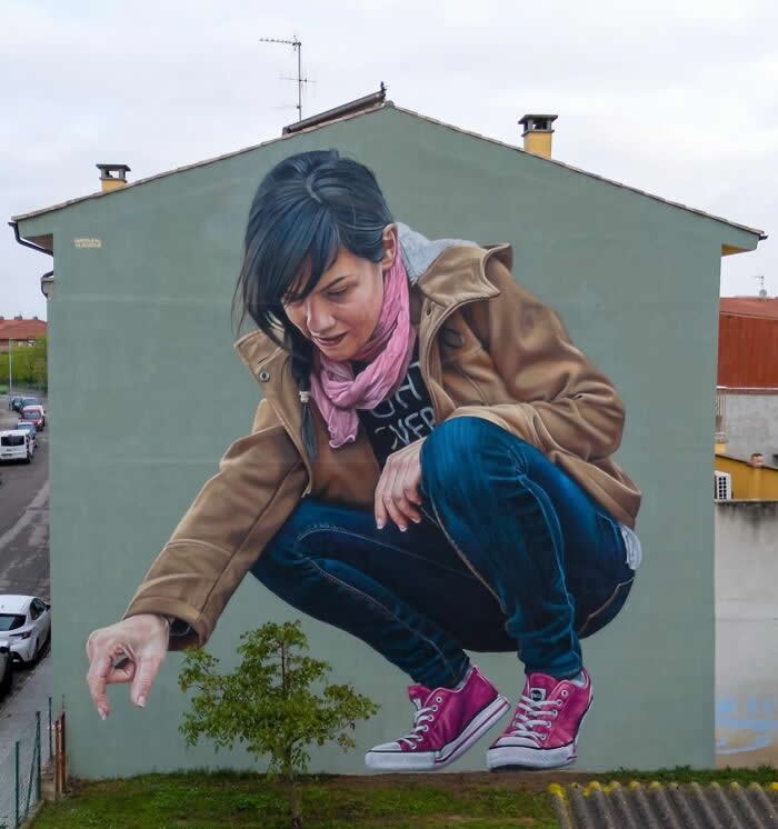 A hyper-realistic mural of a giant woman crouching down and reaching toward a small tree, painted on the side of a residential building, creating a playful illusion of scale in an urban neighborhood. Calldetenes, Spain.