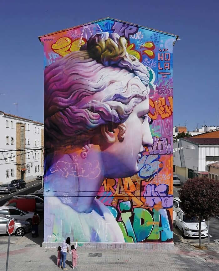 A large mural of a classical sculpture head rendered in soft gradients and vibrant graffiti colors, painted on the side of a building with two people standing below for scale in an urban setting. Mérida, Spain.