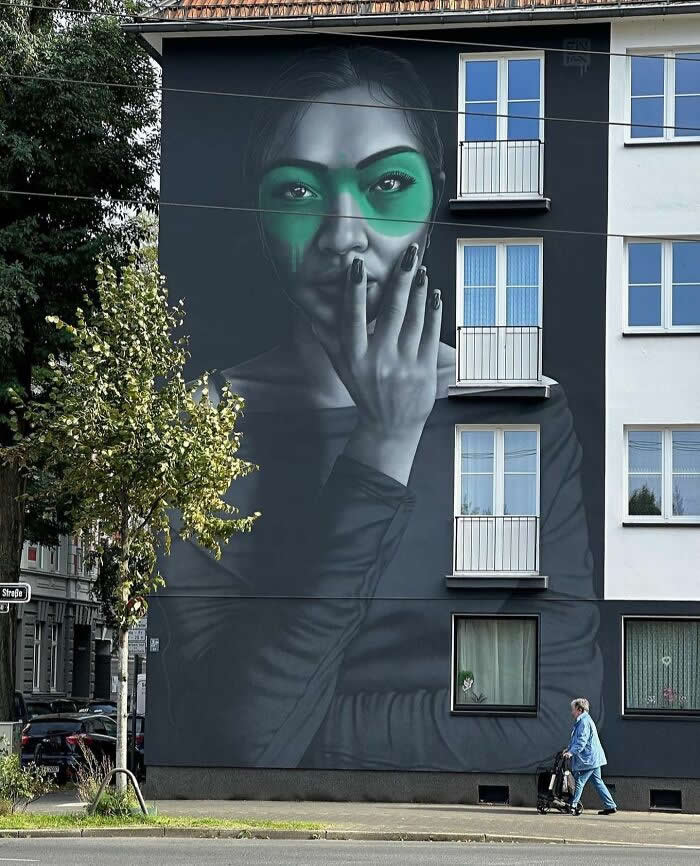 A large grayscale mural of a woman with a green band across her eyes, holding her hand near her face, painted on a building facade with a pedestrian walking past below. Düsseldorf, Germany.