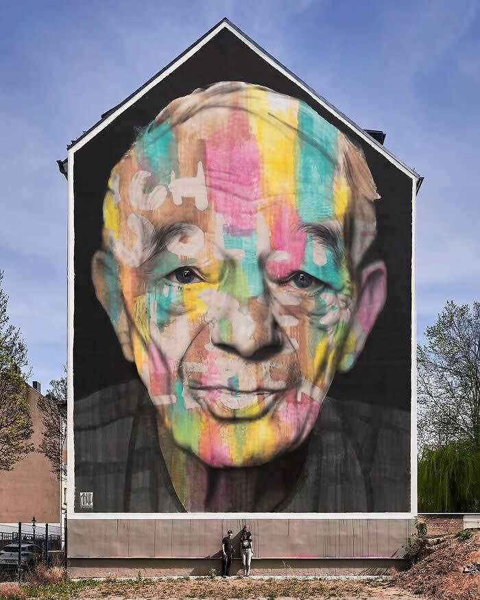 A massive mural of an elderly man’s face layered with colorful abstract paint strokes, covering the side of a building with two people standing below for scale in an outdoor urban setting. Chemnitz, Germany.