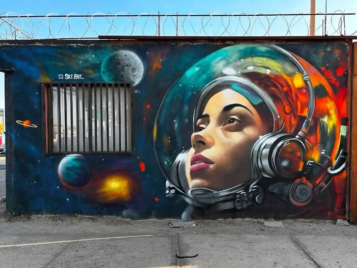 A colorful street mural of a woman wearing a futuristic astronaut helmet, surrounded by planets and space elements, painted on a wall with a barred window integrated into the cosmic scene. Chile.