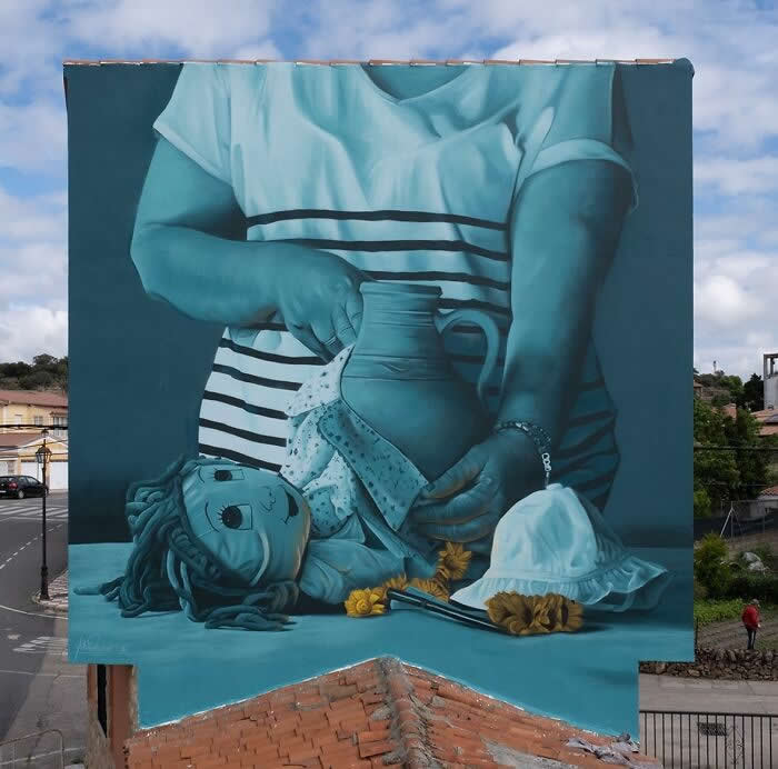 A monochromatic blue mural of a person carefully repairing a broken clay pot, with a doll, flowers, and a hat placed on a table, symbolizing care, nostalgia, and restoration on a building wall. Valdeobispo, Spain.