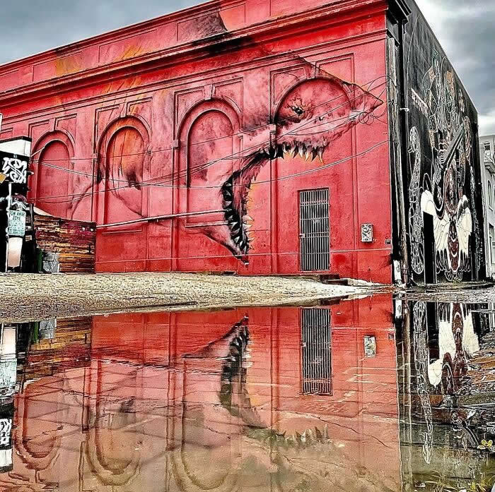 A bold red building mural featuring a surreal creature with a long, jagged mouth emerging across the wall, reflected dramatically in a puddle on the street below in an urban setting. St. Petersburg, Florida, USA.