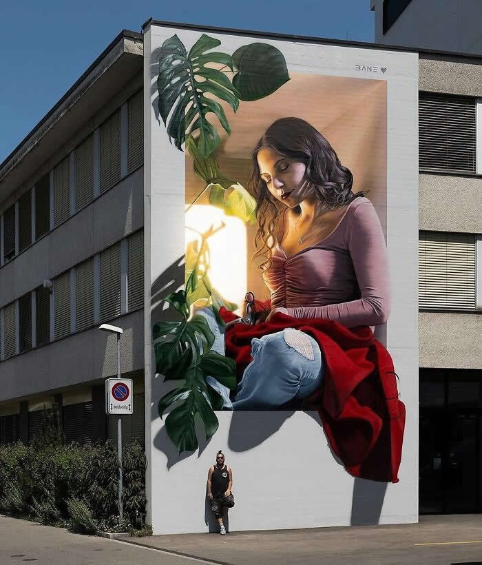 A hyper-realistic mural of a young woman sitting and sewing fabric, surrounded by lush green leaves and warm light, painted on a building wall with a person standing below for scale. Stitching Sunlight and Silence - Frauenfeld, Switzerland.