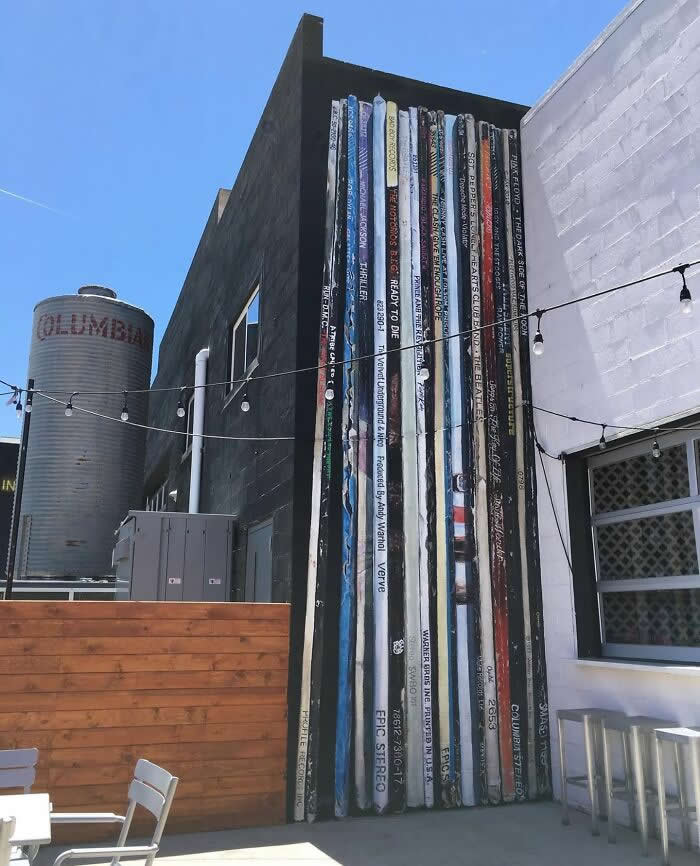 A creative mural of oversized book spines painted along the side of a building, giving the illusion of a towering bookshelf in an outdoor urban courtyard setting. Reno, Nevada, USA.