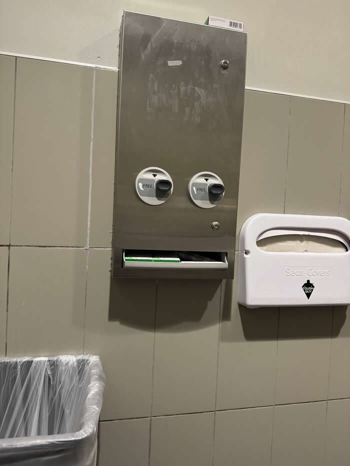 30 Public Objects Having a Total Meltdown 32 A stainless steel restroom wall dispenser with two round knobs that resemble eyes, creating a humorous face-like expression, mounted above a slot for toilet seat covers beside a trash bin and a seat cover dispenser.