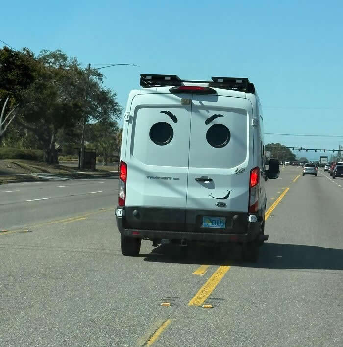 30 Public Objects Having a Total Meltdown 34 The back of a white van on a road with two round window stickers and a curved line forming a playful smiling face, creating a funny expression while driving down a sunny street with trees and cars in the background.