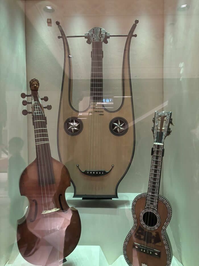 30 Public Objects Having a Total Meltdown 36 A display of musical instruments in a glass case at an opera house museum, where the central guitar has decorative sound holes and a curved bridge forming a smiling face, creating a humorous expression among traditional string instruments.