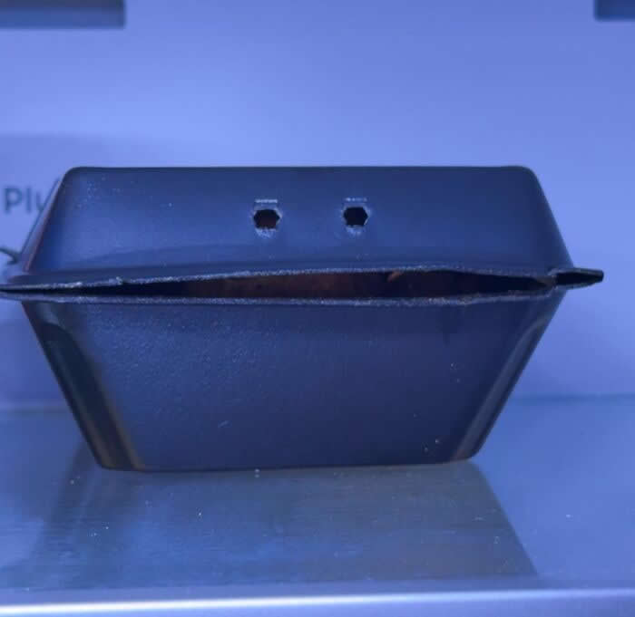 30 Public Objects Having a Total Meltdown 38 A black takeout food container with two small holes resembling eyes and the lid slightly open like a worried mouth, creating a funny face expression while sitting on a counter under blue lighting.