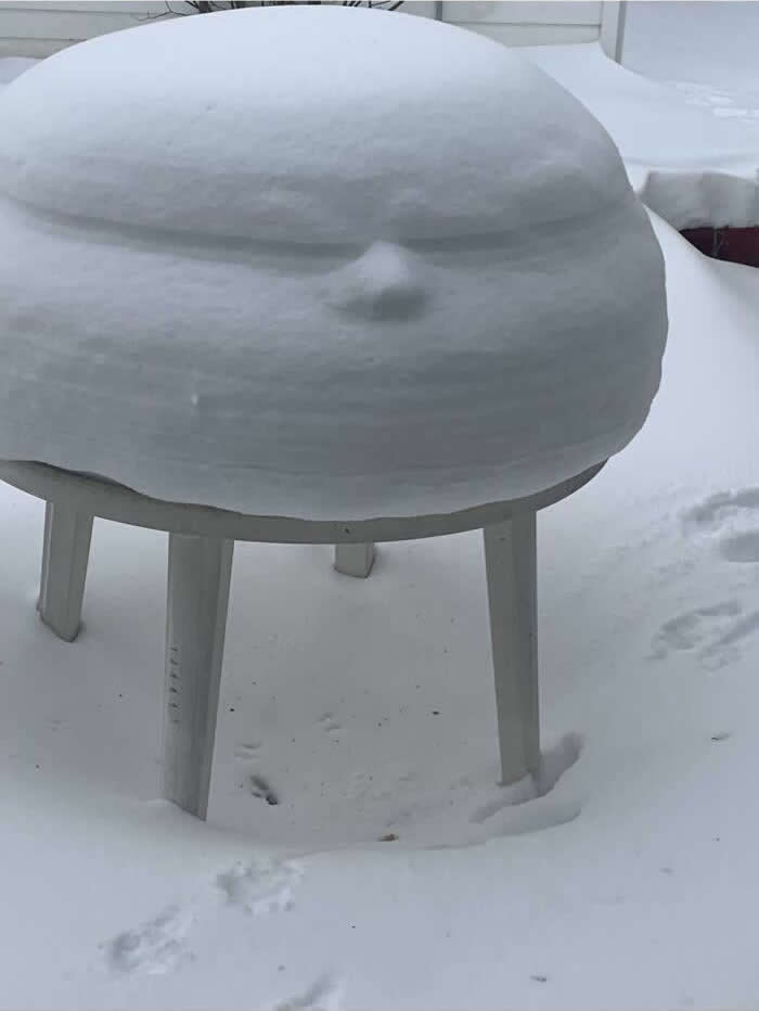 30 Public Objects Having a Total Meltdown 39 An outdoor patio table covered with a thick layer of snow forming a rounded shape with a small bump resembling a nose and a curved line like a grumpy mouth, creating a funny face in a snowy backyard.