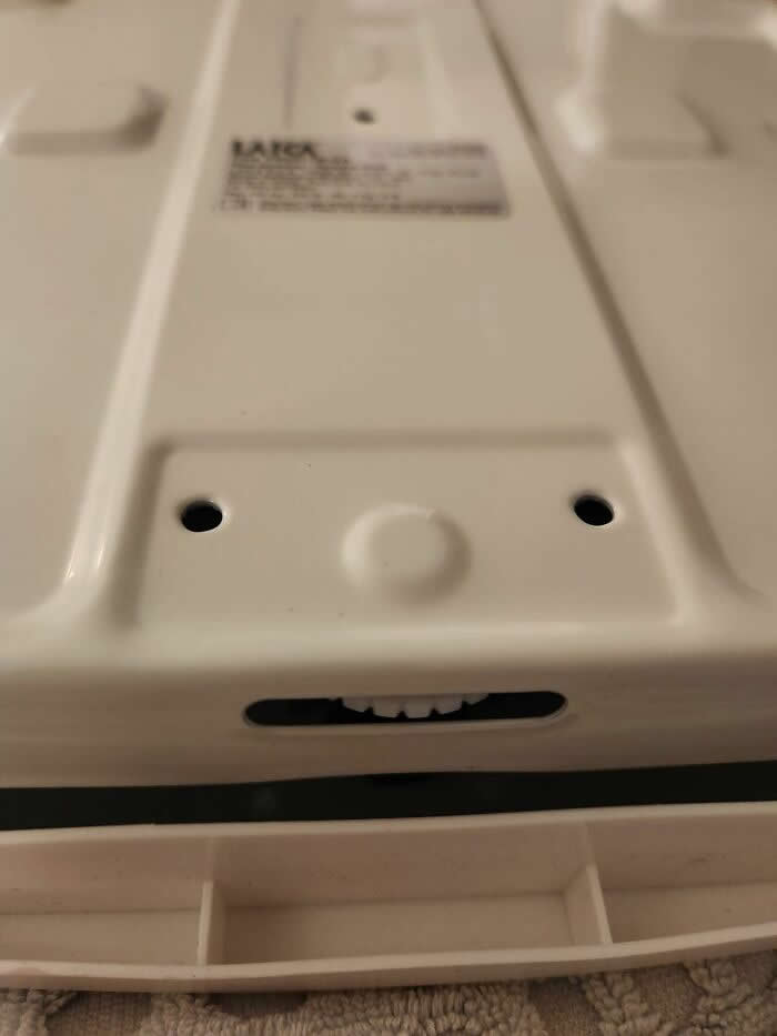 30 Public Objects Having a Total Meltdown 40 The underside of a household appliance with two screw holes resembling eyes, a round bump like a nose, and an opening showing small gear-like shapes that look like teeth, creating a humorous face-like expression.