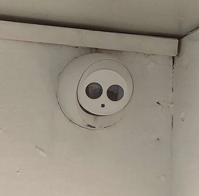 30 Public Objects Having a Total Meltdown 48 A small round security camera mounted in a corner with two dark sensor circles resembling eyes and a tiny hole like a mouth, creating a funny surprised face watching the room.