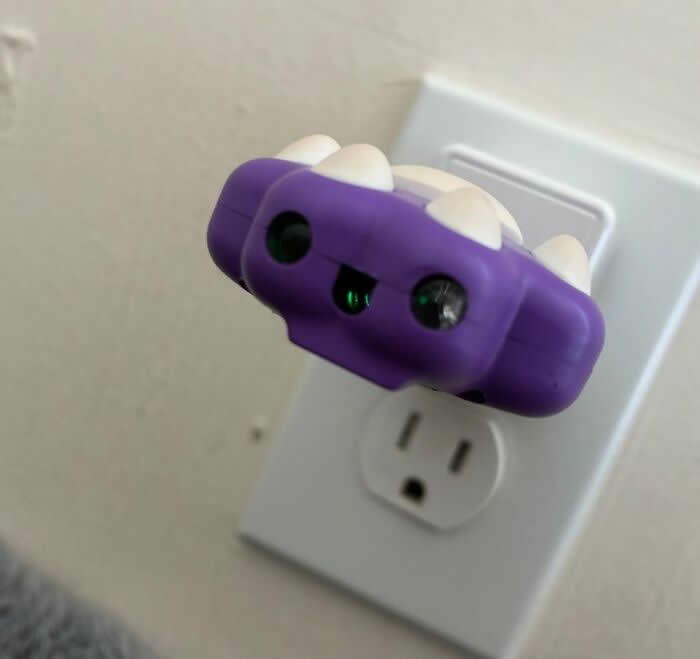 30 Public Objects Having a Total Meltdown 57 A purple laser cat toy plugged into a wall outlet with two glowing lenses resembling eyes and small white spikes like teeth, creating a playful monster-like face ready for playtime.