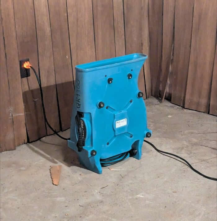30 Public Objects Having a Total Meltdown 58 A blue industrial machine placed on a concrete floor with bolts and shapes on its back forming a smiling face-like expression, creating a funny pareidolia effect against a wooden wall background.