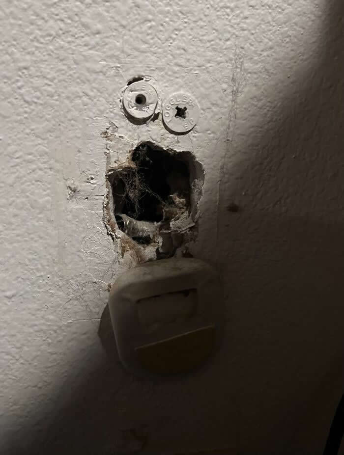 30 Public Objects Having a Total Meltdown 60 A damaged wall outlet area with two white screw caps resembling eyes and a broken hole forming a wide open mouth, creating a surprised face-like expression on the wall through pareidolia.