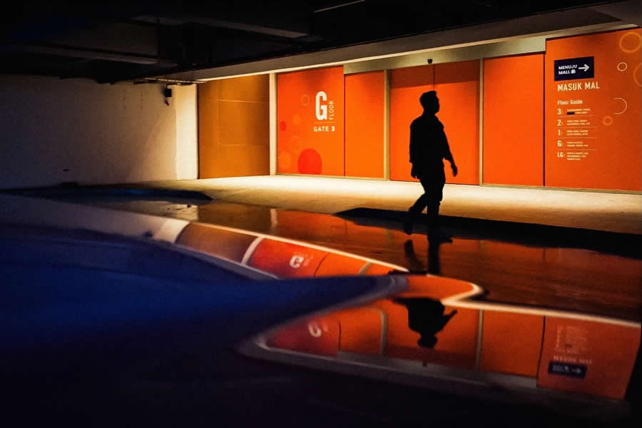 Silhouette of a person walking past a bright orange wall with strong geometric lines, reflected on a glossy surface, showcasing dramatic lighting and contrast in street photography.